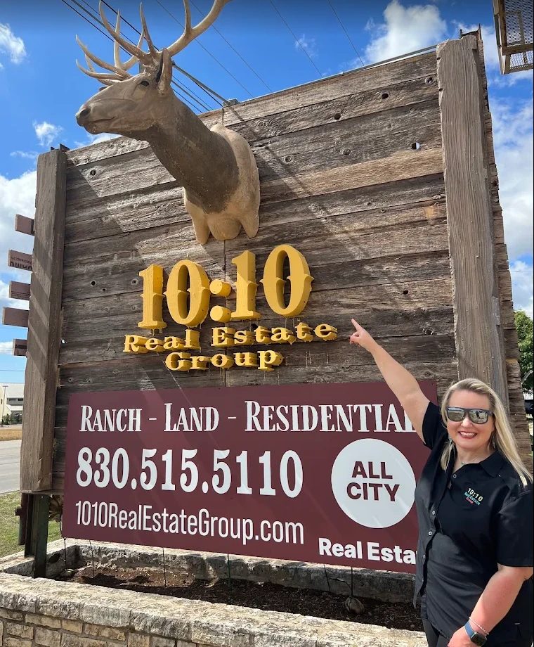 Carrie Farmer pointing at the rustic wooden 10:10 Real Estate Group sign in Kerrville, Texas, with a deer mount above and Ranch, Land, Residential below