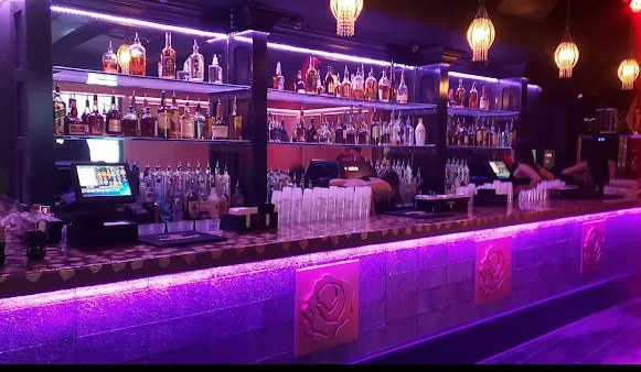 The back bar at Rosebar lit in purple, with shelves of bottles and rose plaques on the front